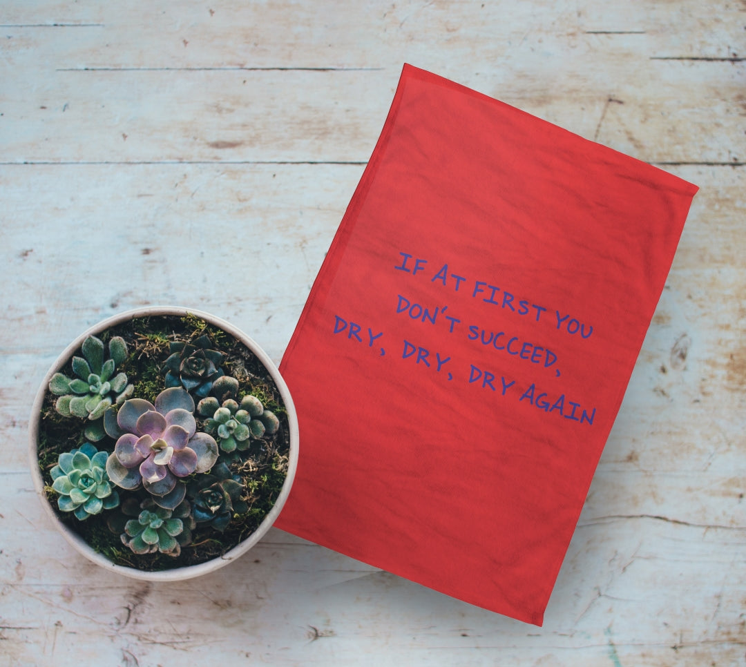 Personalised Red Tea Towel with Text on it. "If At First You Don't Succeed Dry, Dry, Dry Again". On Wooden table next to plant pot.