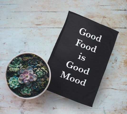 Personalised Tea Towel With Text. Personalised Text is "Good Food is Good Mood". On Wooden Table Next to Plant Pot.