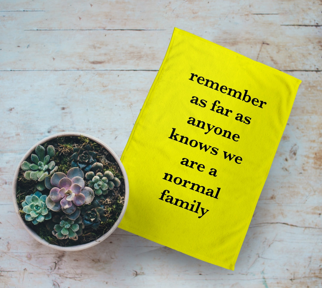 Personalised Yellow Tea Towel with Text on it. "Remember as far as anyone knows we are a normal family". On Wooden table next to plant pot.